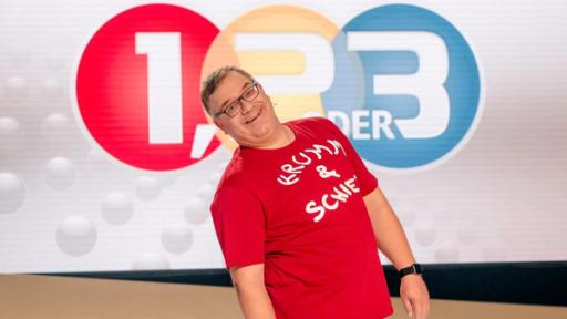 Krumm und schief | Rechte: Ralf Wilschewski 1, 2 oder 3: Moderator Elton ssteht vor der Vidiwall im 1, 2 oder 3-Studio und trägt ein Tshirt mit dem Schriftzug "Krumm und schief".