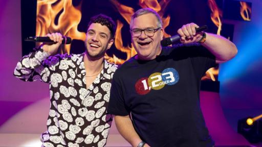 Popstars on Tour | Rechte: ZDF "1, 2 oder 3 - Popstars on Tour": Luca Hänni singt ins Mikrofon. Elton steht dabei. Hinter ihnen sind Flammen zu sehen.