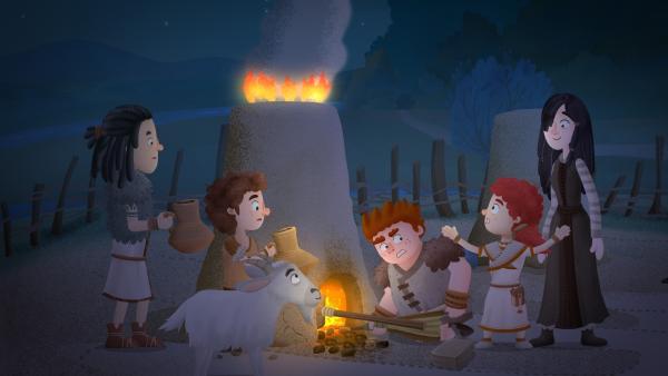 Von links stehen Mimo, Jado, die Ziege, Brock, Leva und Kula vor einem Feuerofen. Brock kniet und hält etwas in das Feuer, Mimo und Jado halten jeweils eine Karaffe.