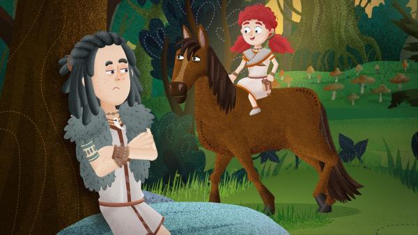 Vorne links Mimo angelehnt an einem Baum. Er blickt grimmig. Im Hintergrund Leva auf einem braunen Pferd und lächelt.