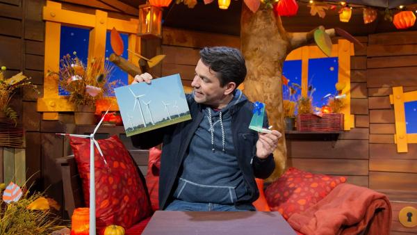 Moderator Juri sitzt im Baumhaus und hält ein Bild von einem Windpark und ein kleines Windrad in den Händen.