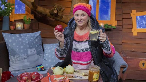 Singa hat in der Hand einen Apfel und ein Teeglas mit heißem Wasser, Apfelschale, Honig und Ingwer.