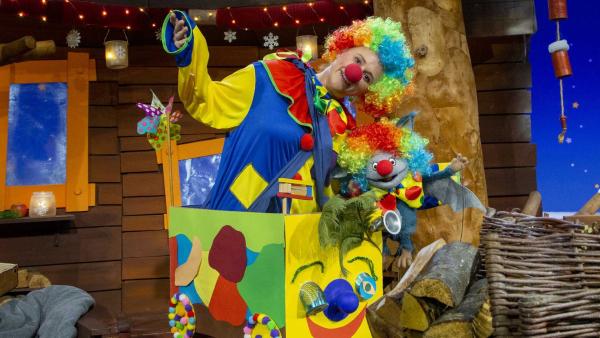 Singa und Fidi tragen beide Clownskostüme und Perrücken in Regenbogenfarben. Sie bringen aus einer riesigen bunten Kiste.