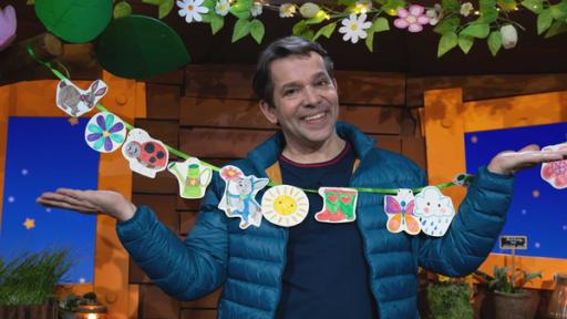 Rechte: KiKA Juri hat die MACH MiT!-Frühlingsgirlande vor sich Hängen. Zu sehen sind auch bunte Gummistiefel.