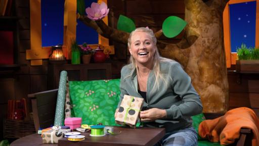 Rechte: KiKA Singa sitzt im Baumhaus. Sie hält ein eingepacktes Geschenk in der Hand und lacht.
