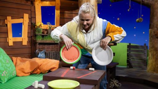 Rechte: KiKA Singa steht mit zwei Papptellern in den Händen vor einem Tisch im Baumhaus, auf dem Korken stehen.
