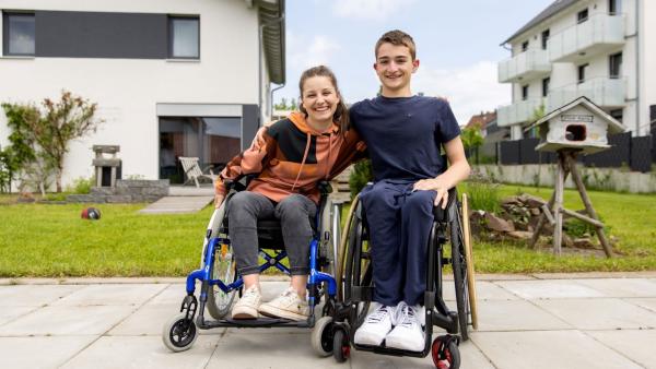Checkerin Marina sitzt links in einem Rollstuhl. Neben ihr ist Rollstuhlfahrer Janis. Er nimmt Marina in den Arm. Beide sitzen draußen auf einer Terrasse.