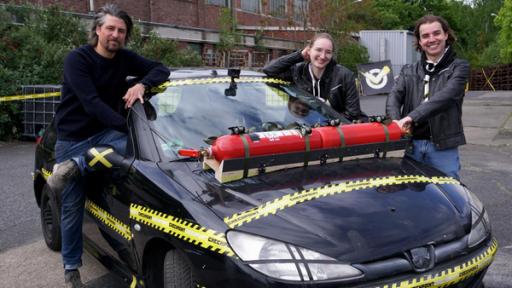 Zwei Jugendliche und ein Mann stehen um ein Auto, auf das Feuerlöscher geklebt sind.