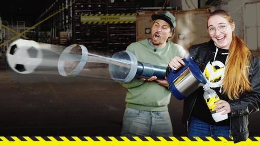In Deckung! Explosive Flaschen und harte Bälle | Rechte: Visual Bridges Checkpoint: Moderator Tommy Scheel schießt aus einer selbst gebastelten Ballkanone einen Fußball ab, während Testteamerin Laura neben ihm steht und dampfende Flüssigkeit in eine Wasserflasche gießt.