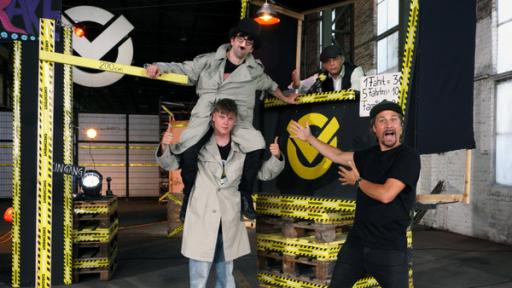 Riesen, Actionhelden und Stapler-Profis | Rechte: ZDF Checkpoint: In der Checkpoint-Halle ist ein Jahrmarktstand aufgebaut, vor dem zwei Testteamer stehen, wobei einer auf den Schultern des anderen sitzt. Moderator Tommy Scheel steht in präsentierender Geste neben ihnen.