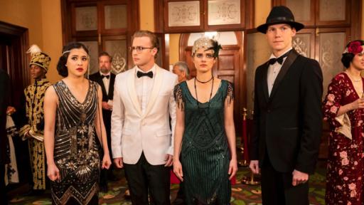Kyra (Kimie Tsukakoshi), Peter (Jamie Carter), Imogen (Elizabeth Cullen) und Darra (Julian Cullen) stehen in festlicher Kleidung in der Mode von 1919 nebeneinander in einer Empfangshalle zwischen anderen Gästen. Sie schauen sich angespannt um.