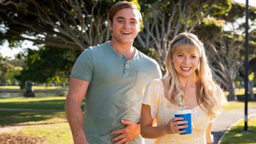 Ben (Matthew Manahan) und Lily (Mia Milnes) spazieren nebeneinander auf einem Weg in einer Parkanlage. Lily hält einen blauen Becher mit einem Strohhalm in der rechten Hand. Beide lächeln glücklich.