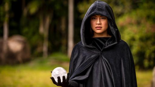 Kyra (Kimie Tsukakoshi) steht, in einen schwarzen Kapuzenumhang gehüllt, in einem Wald. Sie hält in der rechten Hand eine weiße Kugel und blickt ernst nach vorne.