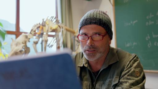 Günther trägt eine Mütze und sitzt im Klassenraum. Er liest aus einem blauen Hefter vor. Dabei schaut er über seine Brille nach rechts.