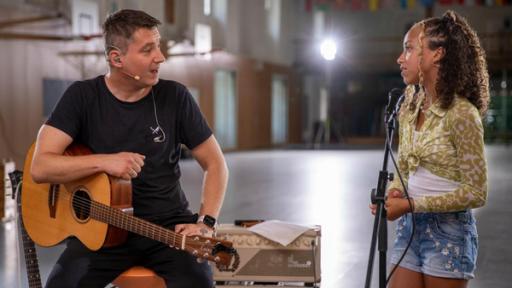 Kandidatin Shayla und Daniel, der Gitarrist der "Dein Song"-Band, stehen nebeneinander und besprechen Shaylas Song. Der Gitarrist sitzt auf einem Hocker, die Gitarre liegt auf seinem Oberschenkel. Shayla steht ihm gegenüber hinter einem Mikrofon und schaut ihn konzentriert an.