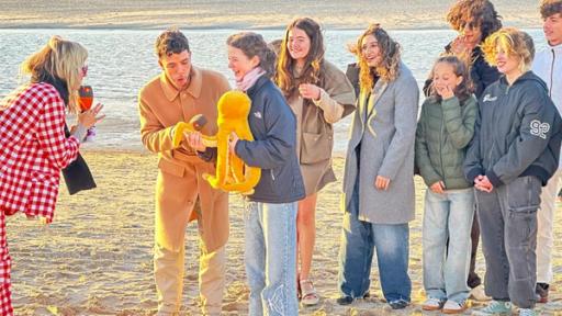 Eine blonde Frau spricht eine Gruppe lachender Jugendlicher an einem Strand an.