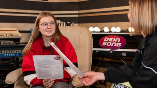 "Dein Song - Gemeinsam kreativ sein - Folge 14": Fine (links) sitzt auf einem Sessel im Tonstudio und hört lächelnd ihrer Patin Mathea zu, die rechts von ihr sitzt. Beide halten Textblätter in der Hand.