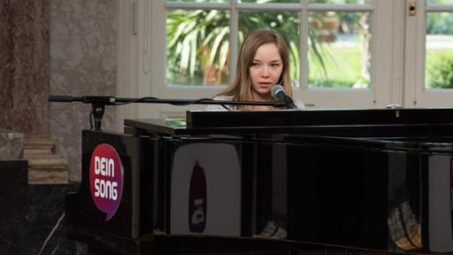 Die "Dein Song" Kandidatin Greta sitzt am Flügel und singt in das Mikrofon. Hinter ihr das Fenster zum Biebricher Schlosspark.