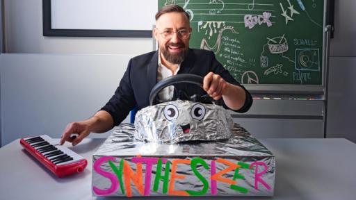 Musikdoktor - Synthesizer | Rechte: ZDF Bürger Lars Dietrich sitz am Schulpult und demonstriert vor einer einer bunt beklebten Kiste mit Lenkrad, wie ein Synthesizer funktioniert.