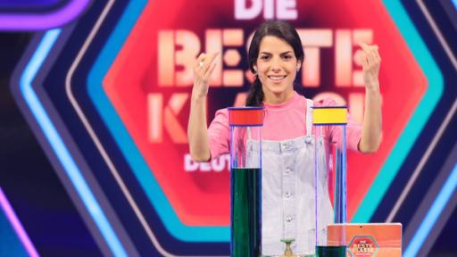 Rechte: KiKA/Steffen Becker Im Studio. Clarissa lächelnd vor dem Logo "Die beste Klasse Deutschlands". Vor ihr auf einem Tisch zwei große Reagenzgläser, die mit grüner Flüssigkeit gefüllt sind.