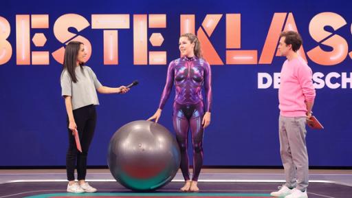 Rechte: KiKA/Steffen Becker Im Studio: In der Mitte “Pezziball-Artistin“ Monique Schröder im Sportdress und einem großen Pezziball. Ihr Gesicht ist Clarissa zugewandt, die links neben ihr steht. Rechts steht Tobi und schaut zu ihnen.