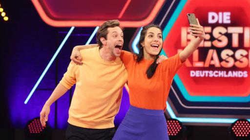 Rechte: KiKA/Steffen Becker Clarissa hat ihren Arm um Tobi gelegt und macht aus der Bewegung heraus ein Selfie mit dem Smartphone. Beide lachen mit weit geöffnetem Mund. Im Hintergrund die Studiokulisse mit dem Logo von "Die beste Klasse Deutschlands".