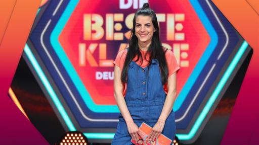 Rechte: KiKA/Steffen Becker Clarissa steht mittig und lächelt. In ihren Händen hält sie eine Moderationskarte. Im Hintergrund das Logo von Die beste Klasse Deutschlands.