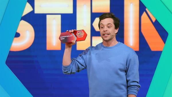 Mittig steht Tobi. Er hält in seiner Hand ein kleines Schild aus Plastik mit der Aufschrift Superfinalist 2024 und dem Logo von Die beste Klasse Deutschlands. Im Hintergrund große Buchstaben.