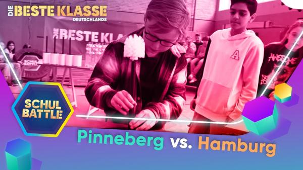 In der Mitte ein Schüler von vorne an einem Tisch. Mit gesenktem Kopf hält er mit beiden Händen eine Klobürste mit der Bürste nach oben. Rechts neben ihm steht ein Schüler, der zu ihm schaut. Im Hintergrund weitere Kinder, die zuschauen.