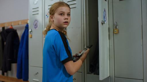 Malin hat ein blaues T-Shirt an und hält ein Handy in der Hand. Sie steht vor grauen Schulspinten.  