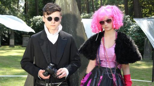 Der verschwundene Engel | Rechte: NDR/Studio Hamburg Foto: Romano Ruhnau Ein Junge (Tom Böttcher) und ein Mädchen (Johanna Werner) mit Sonnenbrillen und auffälliger Kleidung stehen unter Bäumen.