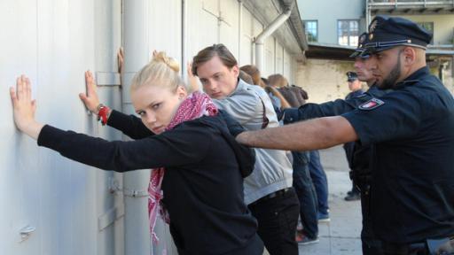 Eine junge Frau (Eva Nürnberg) und ein junger Mann (Alexander Finkenwirth) stehen mit anderen jungen Menschen mit den Händen an einer Wand. Hinter ihnen stehen Polizisten.