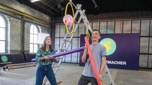 Golf, Baumklettern und Tennis | Rechte: Annalena Renneisen Die Sportmacher: Laura und Gastmoderator Eric Mayer in der Sportmacher-Halle. Beide halten eine Schwimmnudel in der Hand. Im Hintergrund steht eine große Leiter und das Sportmacher-Logo.