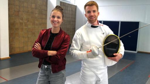 Die Sportmacher: Moderner Fünfkampf | Rechte: Annalena Renneisen Die Sportmacher: Die Moderatoren Laura und Stefan stehen in der Turnhalle. Stefan trägt einen Fechtanzug und hält ein Florett in der Hand. Unter dem Arm trägt er eine Schutzhaube. Laura steht lachend mit verschränkten Armen neben ihm.