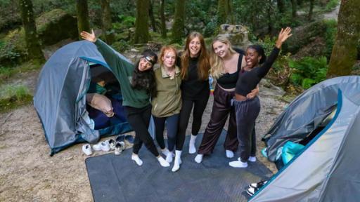 MWG Porto - Campen in der Wildnis | Rechte: ZDF, E+U TV Fünf Mädchen stehen vor zwei Iglu-Zelten in einem Wald. Sie tragen Jogginghosen. Die Zelte sind offen, es liegen Schlafsäcke und Luftmatratzen darin. Vor den Zelten liegen die Turnschuhe der Mädchen. Die Fünf haben die Arme umeinander gelegt und lachen in die Kamera.