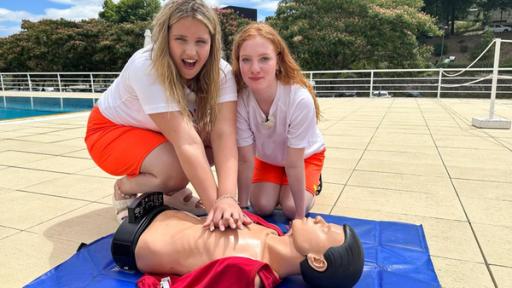 MWG Porto - Die Rettungsschwimmerinnen | Rechte: ZDF, E+U TV Zwei Mädchen üben anhand eines Puppen-Torsos die Herzdruckmassage. Beide Mädchen tragen weiße T-Shirts und orangefarbene Shorts. Das blonde Mädchen hält die Hände übereinander auf der Brust des Puppen-Torsos, um dort die Erste-Hilfe-Maßnahme zu üben. Neben ihr kniet ein rothaariges Mädchen und lächelt in die Kamera.