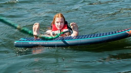MWG Porto - Mit Speed hinterm Boot | Rechte: ZDF, E+U TV Mitten im Waser liegt ein Waveboard. Dahinter ist der Kopf eine rothaarigen Mädchens zu sehen, sie hält in der Hand eine grüne Zugleine. Die Füße streckt sie nach vorne in die Kamera, die Fersen liegen an der Kante des Boards auf.