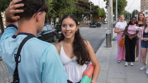 MWG Porto - Nachhilfe im Flirten | Rechte: ZDF, E+U TV Mit dem Rücken zur Kamera steht ein Junge, der sich verlegen den Hinterkopf hält. Vor ihm steht ein Mädchen mit langen dunkelbraunen Haaren und lacht ihn an. Offenbar stehen die beiden an einer Bushaltestelle. Im Hintergrund links fahren Autos auf der Straße, rechts im Bild stehen vier weitere Mädchen und beobachten die Szene lachend.