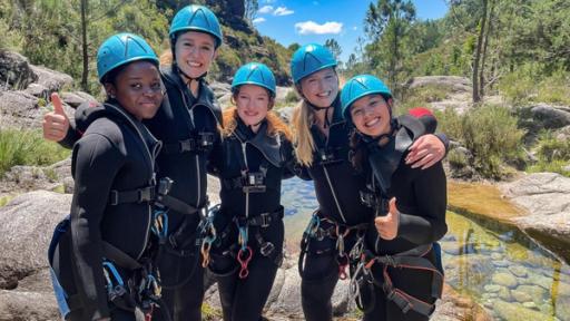 MWG Porto - Team Adrenalin | Rechte: ZDF, E+U TV Fünf Mädchen in Neoprenanzügen mit blauen Helmen auf dem Kopf und Sicherungsgurten um den Körper stehen an einem Fluss mit großen Steinen. Sie haben die Arme umeinander gelegt, zwei Mädchen recken den Daumen in die Höhe.