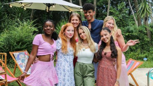 MWG Porto - Videodreh mit KATI K und Lockerlouis | Rechte: ZDF, E+U TV Sechs jugendliche Mädchen und ein Junge stehen als Gruppe in einem Garten. Im Hintergrund sind Liegestühle und ein Sonnenschirm zu sehen. Alle haben die Arme umeinander gelegt und lächeln in die Kamera.