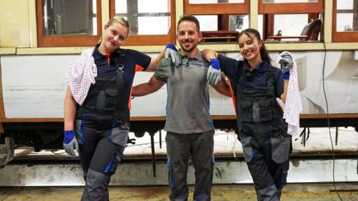 MWG Porto - Zeitreise mit Ölwechsel | Rechte: ZDF, E+U TV Zwei Mädchen haben sich auf die Schultern eines jüngeren Mannes gestützt, er legt freundschaftlich die Arme an ihre Rücken. Die Mädchen tragen blaue Arbeitsoveralls und grau-blaue Arbeitshandschuhe. Das dunkelhaarige Mädchen auf der rechten Seite des Fotos hält ein weißes Tuch mit roten Punkten locker in die Höhe, das blonde Mädchen auf der linken Seite hat sich so ein Tuch über die rechte Schulter geworfen. Im Hintergrund ist eine Trambahn zu sehen. Es handelt sich um eine Tram-Werkstatt in Porto, Portugal.