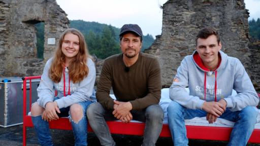 Draußen schlafen Auf der Burg Teaserbild neu | Rechte: ZDF Tommy sitzt mit zwei Jugendlichen vom Draußen schlafen-Team auf einem roten Metallbett. Alle schauen erwartungsvoll in die Kamera.