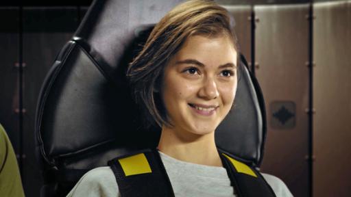 Außer Kontrolle | Rechte: Sklan&Ka/NDR Groß im Bild ist Astronautin Emma glücklich lächelnd, angeschnallt im Sitz des Beschleunigungs-Simulators.