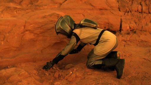 Verrat | Rechte: Sklan&Ka/NDR Eine Astronautin kniet seitlich unter einem großen Felsen und verbuddelt etwas mit den Händen.