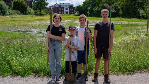 Vier Kinder stehen am Straßenrand vor einer grünen Wiese mit Gartenwerkzeugen in der Hand.