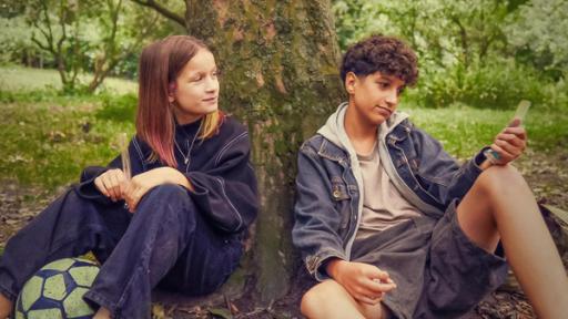 Echt Friends - Teaser - Folge 4 | Rechte: © ZDF / Studio Zentral Liv und Pit sitzen im Grünen unter einem Baum. Beide haben ein Eis in einer schmalen durchsichtigen Tüte in der Hand. Vor Liv liegt ein Fußball. Liv blickt lächelnd und ein bisschen erwartungsvoll zu Pit, der leicht lächelnd nach vorne auf sein Eis schaut.