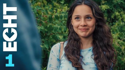 Leyla (Aleyna Kuş) lächelt verlegen aber auch erwartungsvoll jemanden an, der ihr gegenüber steht. Sie steht draußen, im Hintergrund eine Hecke.