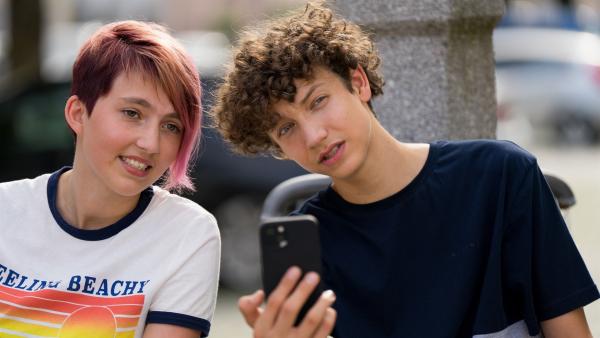 Carli und Edvard nah nebeneinander. Edvard hält ein Smartphone vor sich in der Hand und schaut prüfend auf etwas in der Ferne.