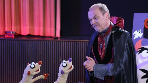 Ganz schön gruselig – Teil 3 | Rechte: bigSmile/NDR Zwei Erdmännchen gucken einen Mann mit Umhang an. Im Hintergrund ist ein roter Vorhang und eine Bühne.