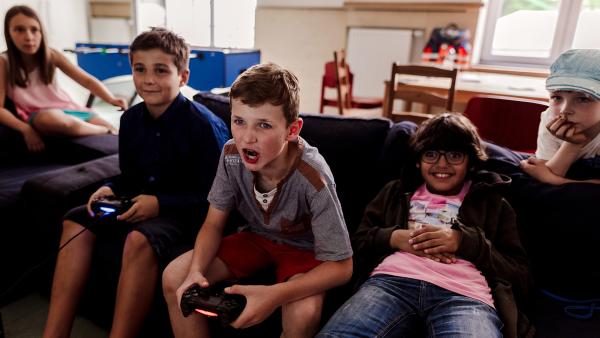 Vier Jungen sitzen auf einer Couch und spielen mit einer Konsole. Ein Mädchen im Hintergrund schaut zu.